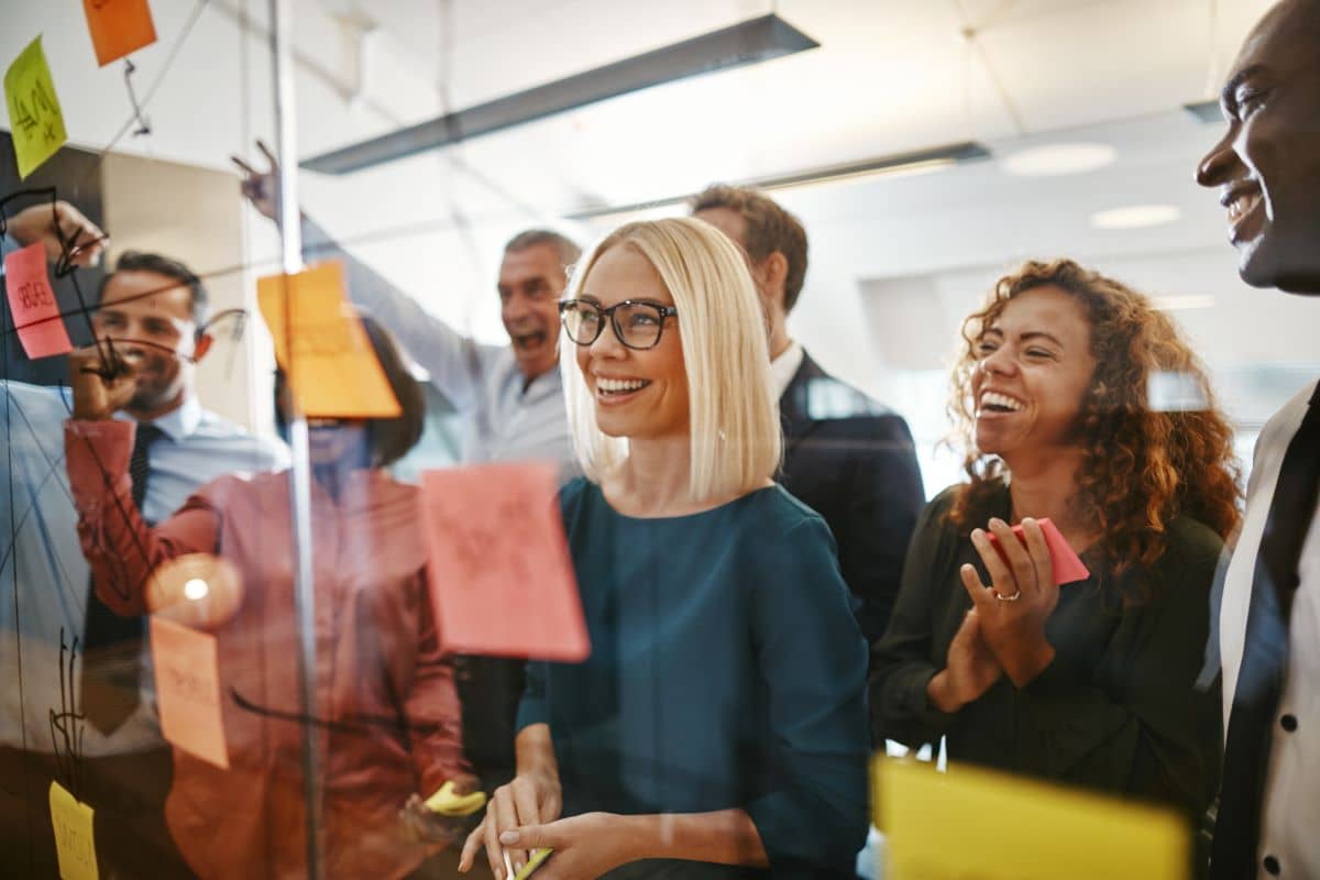 This is a picture of a group of coworkers standing and facing a large glass wall that has post-it notes on it. It appears that the team is having a brainstorm session.