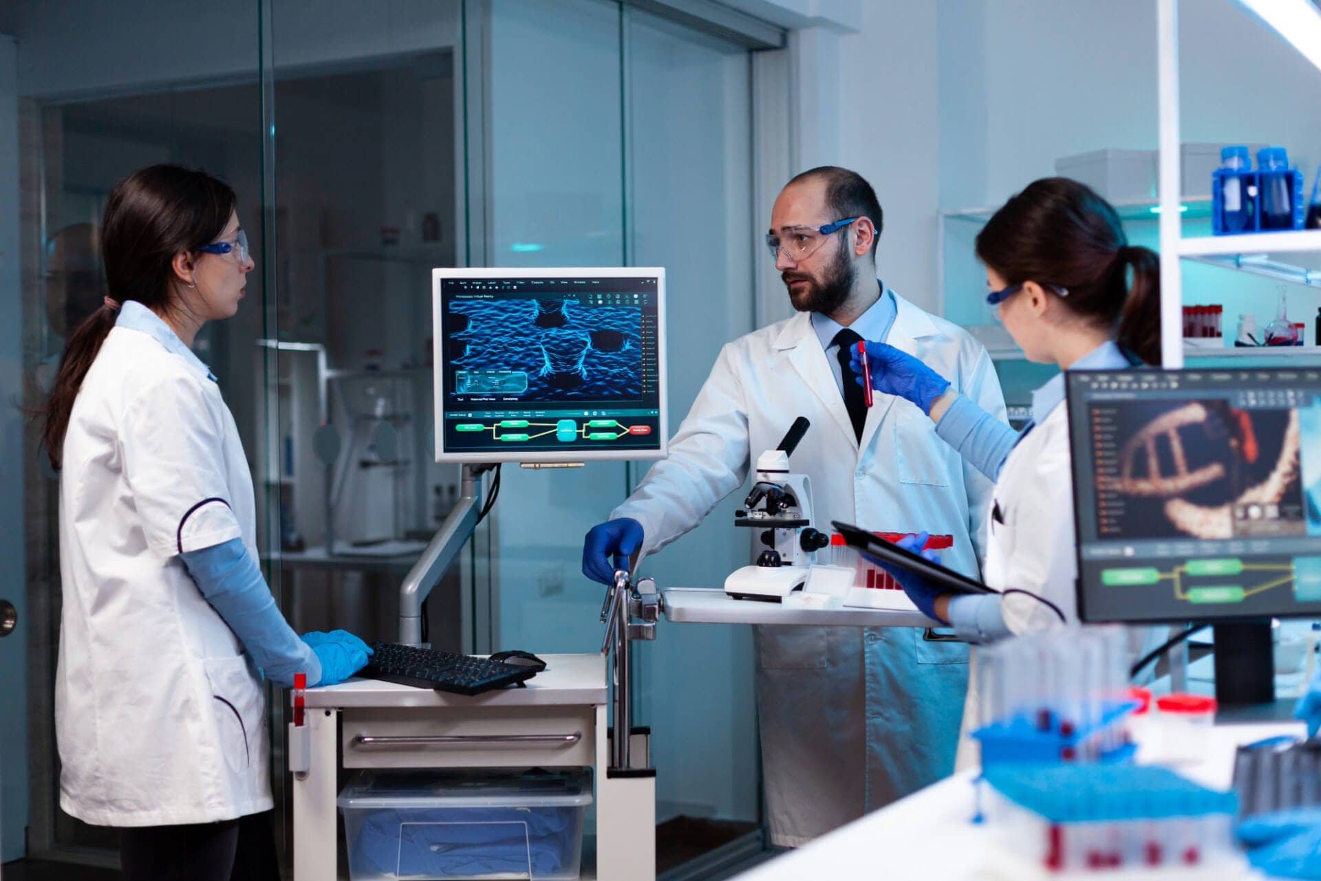 Three laboratory professionals stand in a lab around a computer display screen. All three wear lab coats. The man is speaking while two female laboratory scientists look at him.