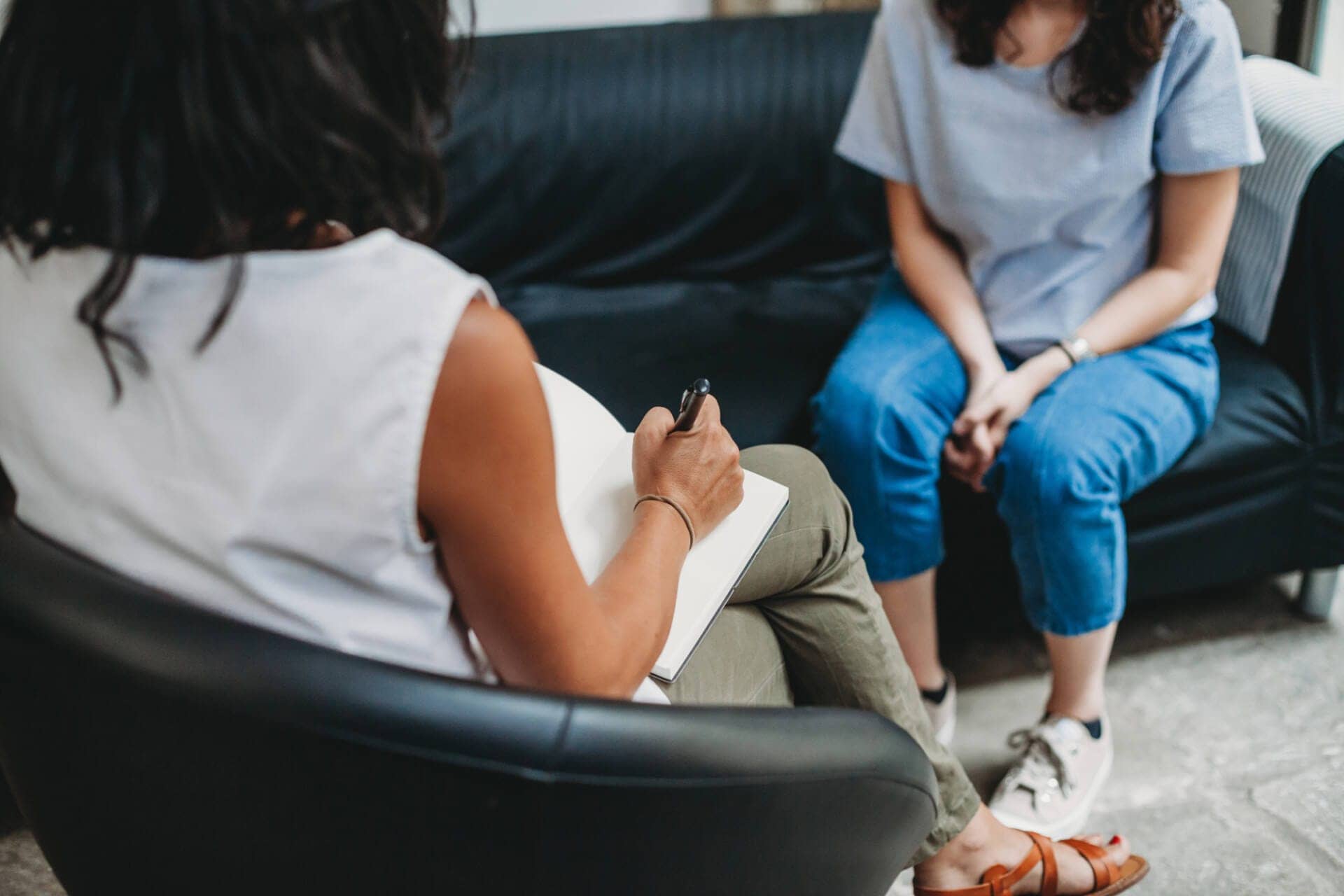 A female mental health professional sits in a chair and writes in a notebook. A woman in jeans and a t-shirt sits on the couch across from her, hands folded in her lap.
