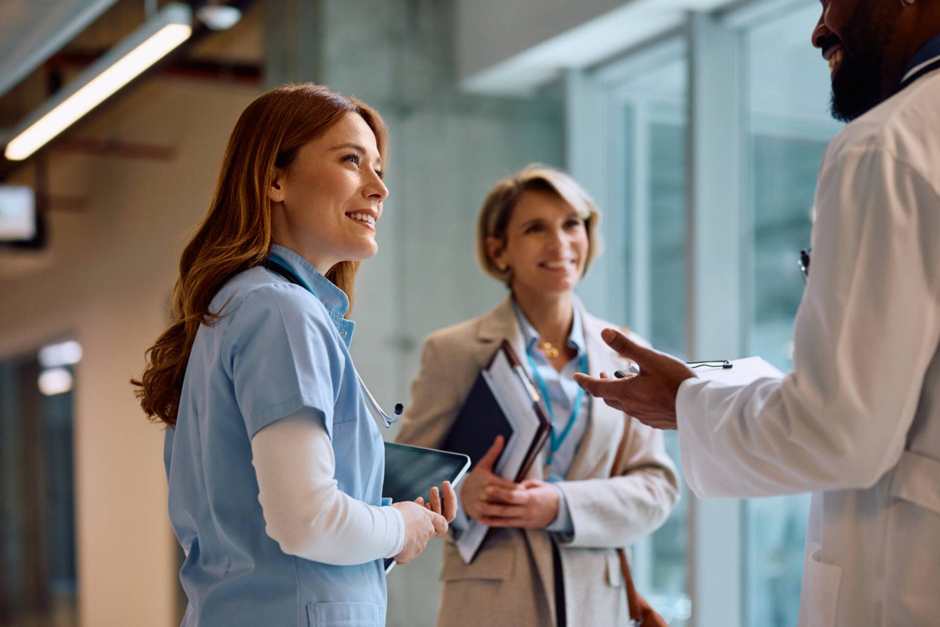 A healthcare administrator in a tan blazer talks to a male doctor off screen and a female nurse with long red hair and blue scrubs. They smile at each other with a glass door and hospital setting in the background.