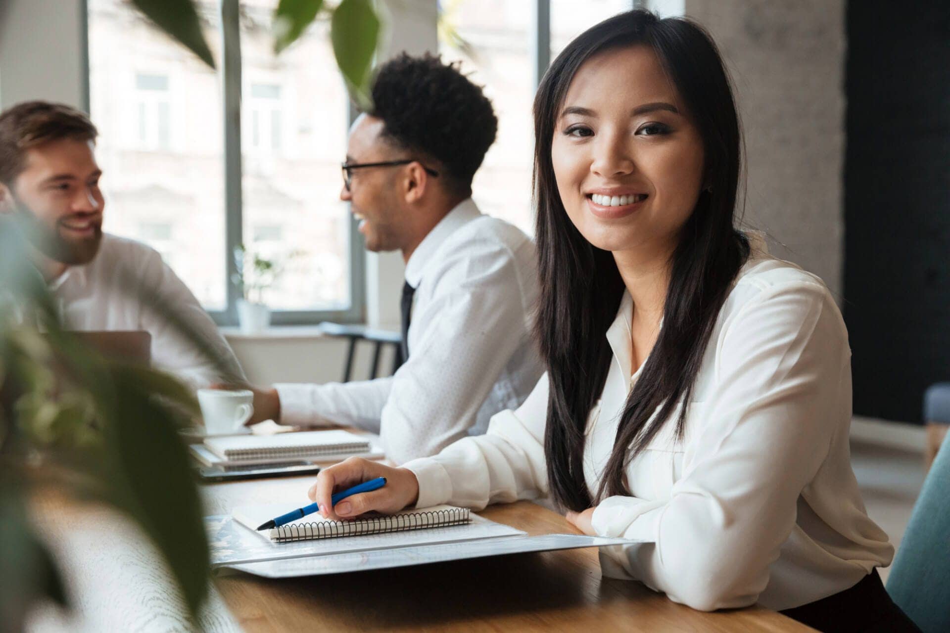 A female paralegal professional sits and smiles. She is wearing a white blouse and sitting at a table. She holds a blue pen on top of a notebook and file folder. Behind her, two men in white button-down shirts are talking at the table.