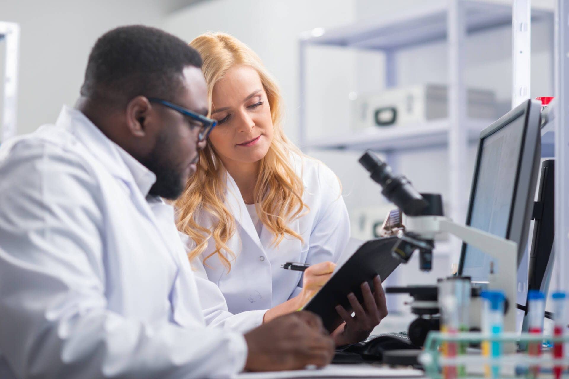 A man and woman sit in front of a microscope and a computer in a clinical laboratory setting. The man holds a clipboard that the woman makes a note on with a pen.
