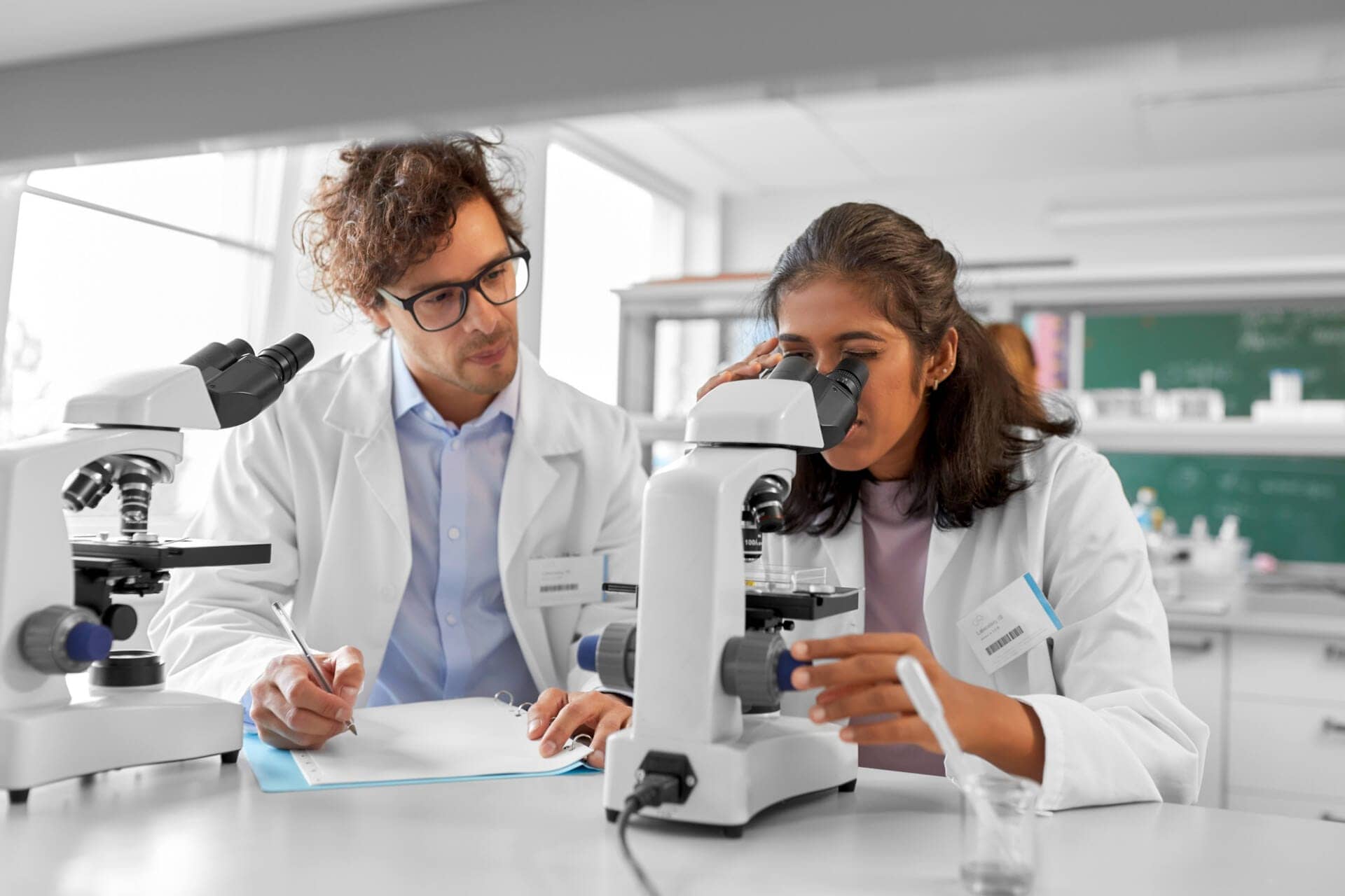 A male DCLS professional provides guidance to a female clinical laboratory professional. They are both wearing lab coats. The woman has shoulder length brown hair and is looking into a microscope. The man is writing on a paper on the table.