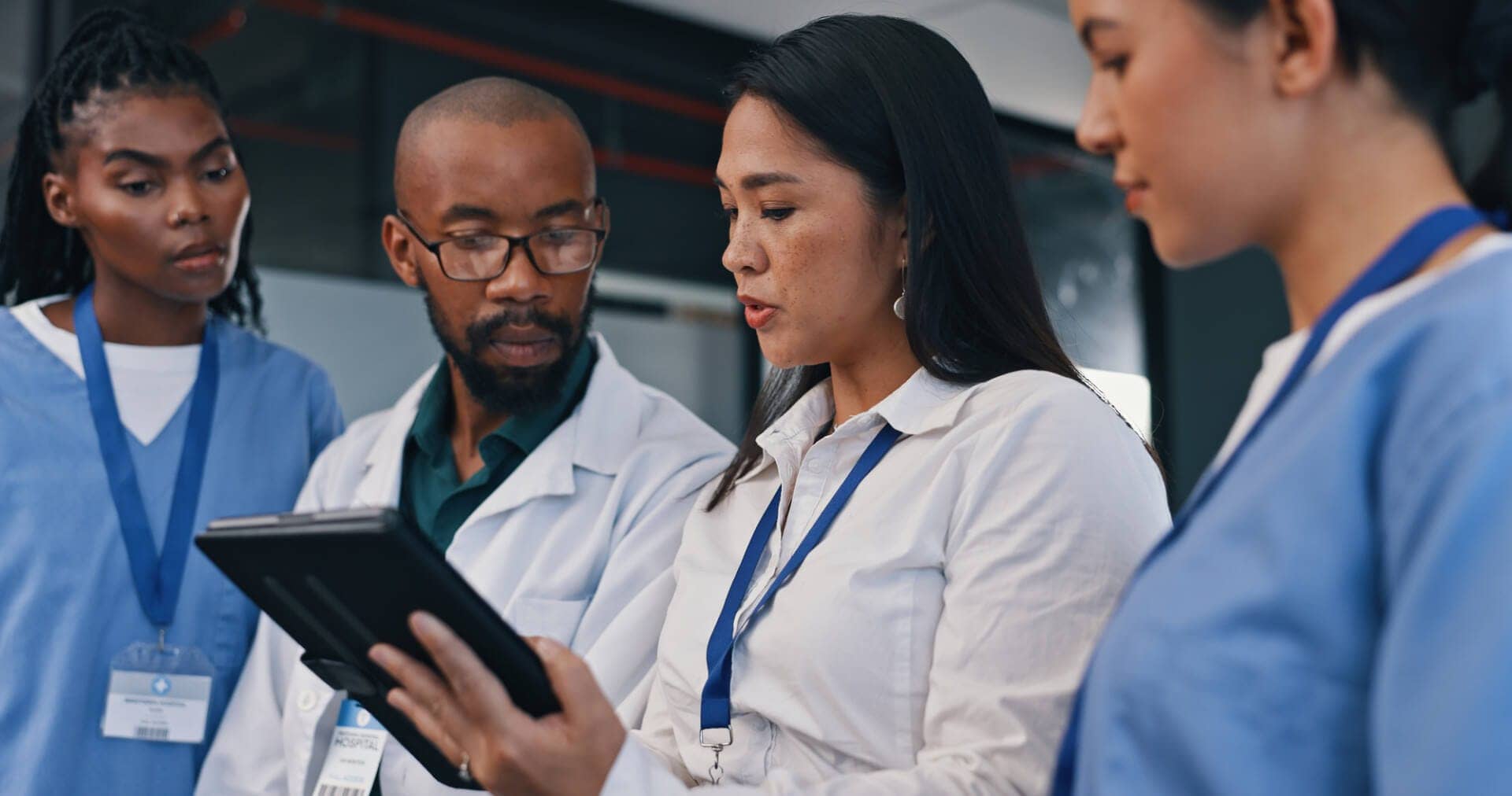 A female health informatics professional wears a white shirt and holds a patient records folder. She is surrounded by a male doctor wearing a white lab coat and two nurses wearing blue scrubs. They are listening to her explain the patient records in her hands.