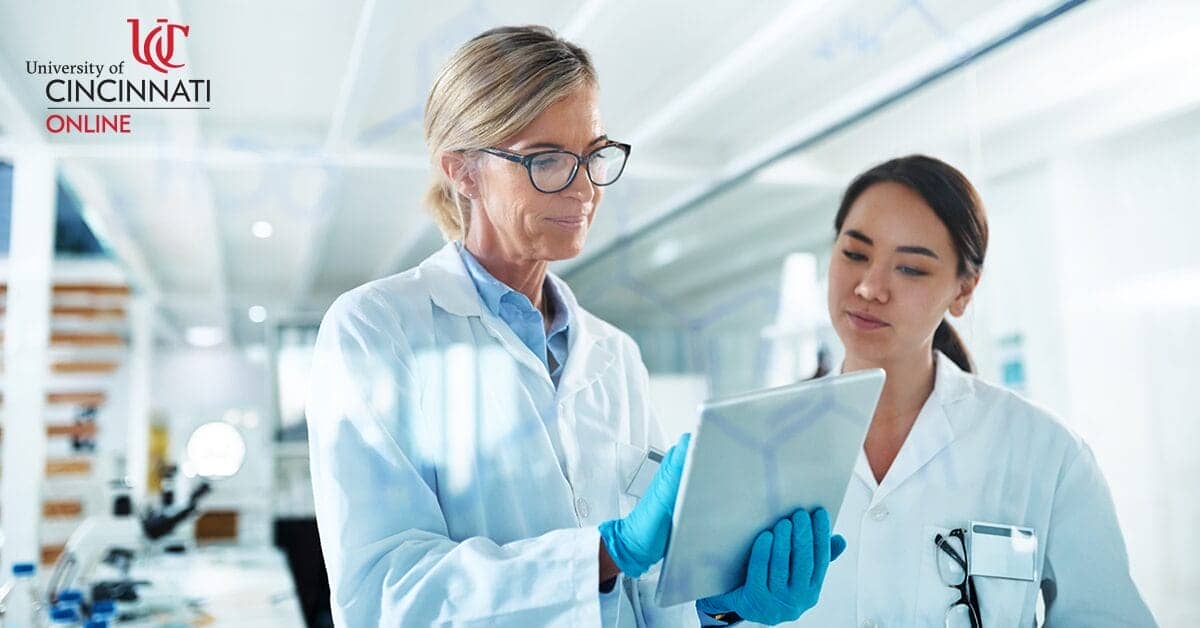 Two women stand in a laboratory in white lab coats. The woman on the left is older; she has blonde hair and wears glasses. The other woman is younger with dark hair. The woman on the left holds a clip board and gestures to it while the other woman looks.