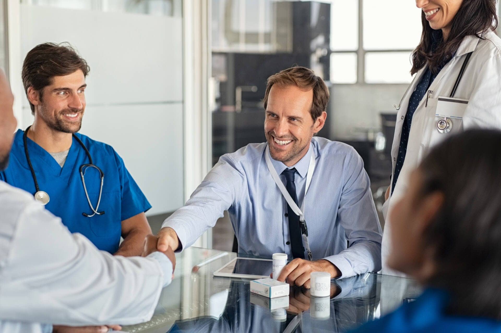 A male healthcare operational excellence (HOPEX) professional sits at a table. He wears a blue button down shirt and tie, and he is smiling as he shakes hands with a man on the other side of the table. He is surrounded by doctors and nurses who are all smiling. 