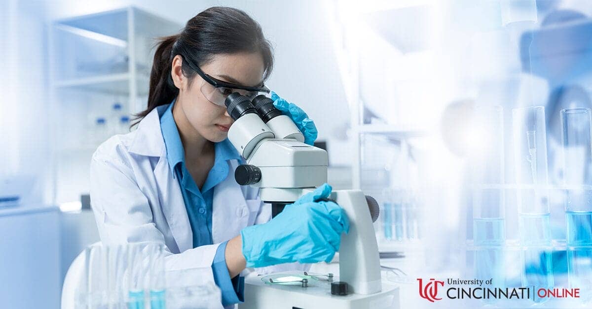A woman with a dark ponytail wears a white lab coat and laboratory eye wear as she looks into a microscope. She is in a bright clinical laboratory setting.