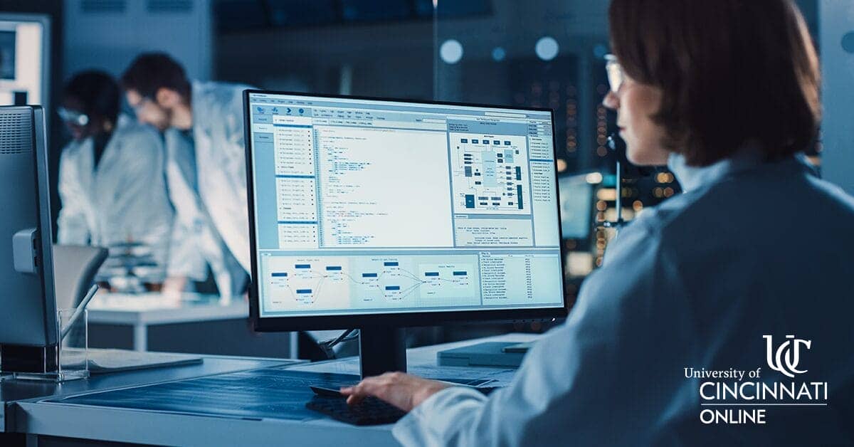 A woman with short brown hair sits at a desk, facing away from the camera. She is looking at medical data on a computer. Two lab professionals are leaning over a table in the background, out of focus.