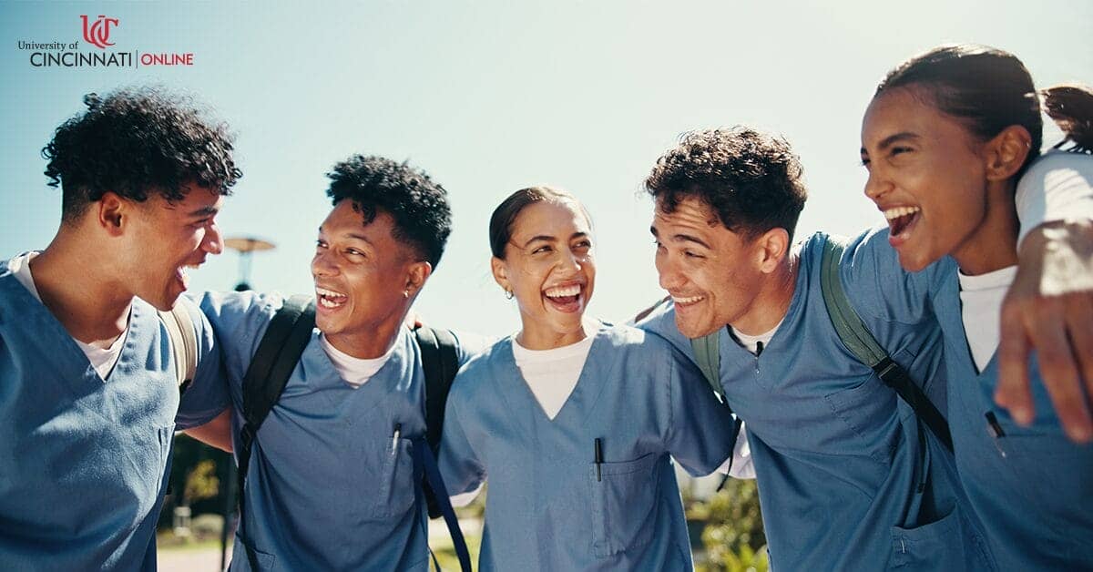 A group of nurses stand in a semi-circle with their arms over each other's shoulders. They are all in blue scrubs and laughing as they smile at one another.