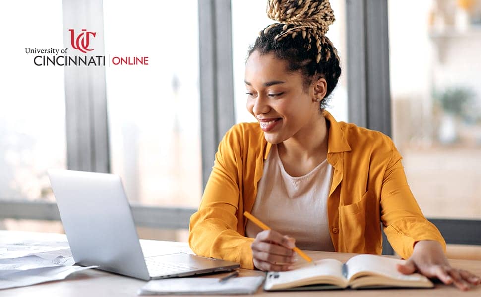 A woman in a yellow shirt with a high bun smiles at her open laptop. She holds a pencil and writes in a notebook at a desk. There are windows behind her.