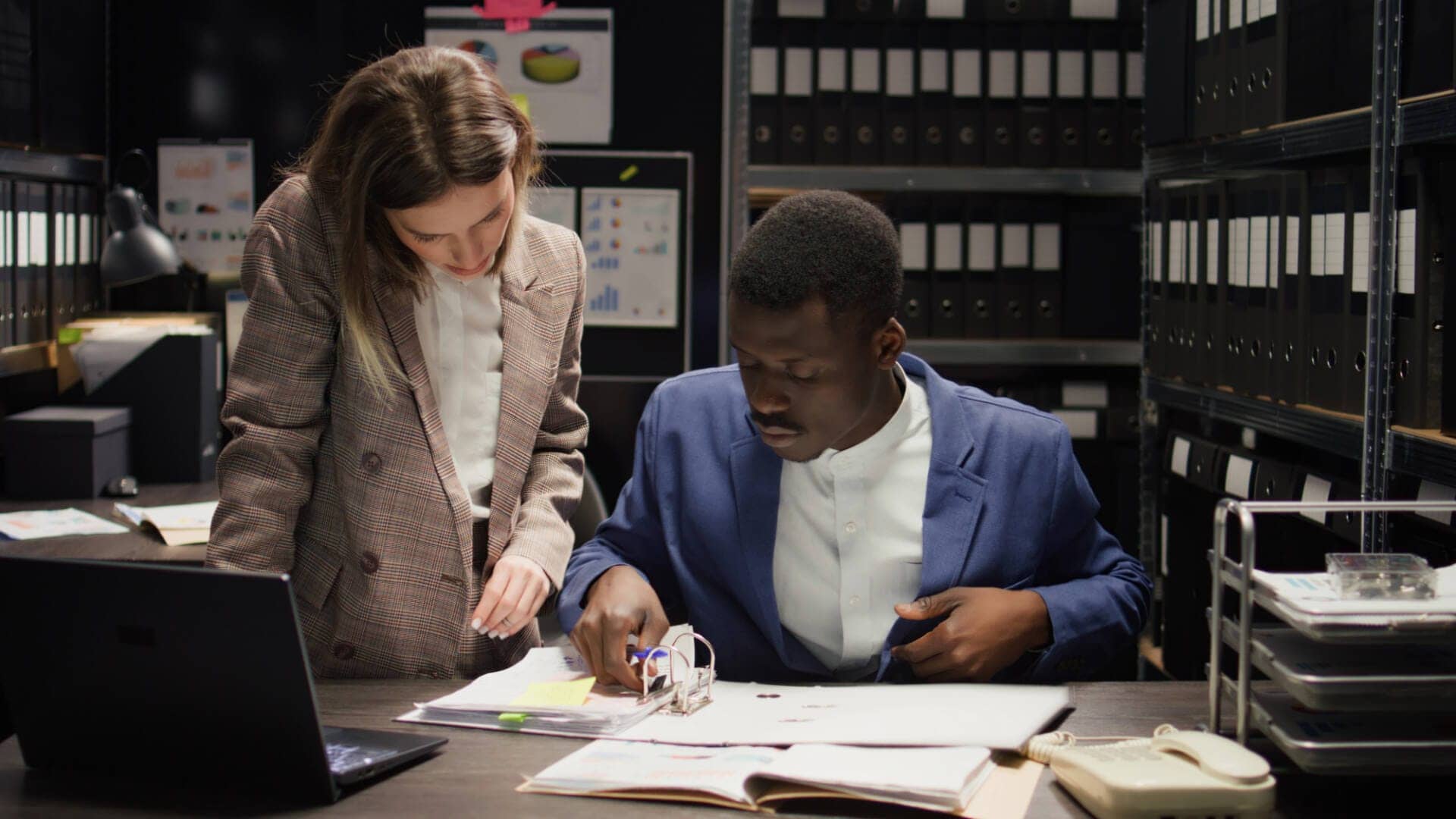 Two private detectives lean over a table covered with papers. The man sits at the table while the woman is standing and looking down at it.