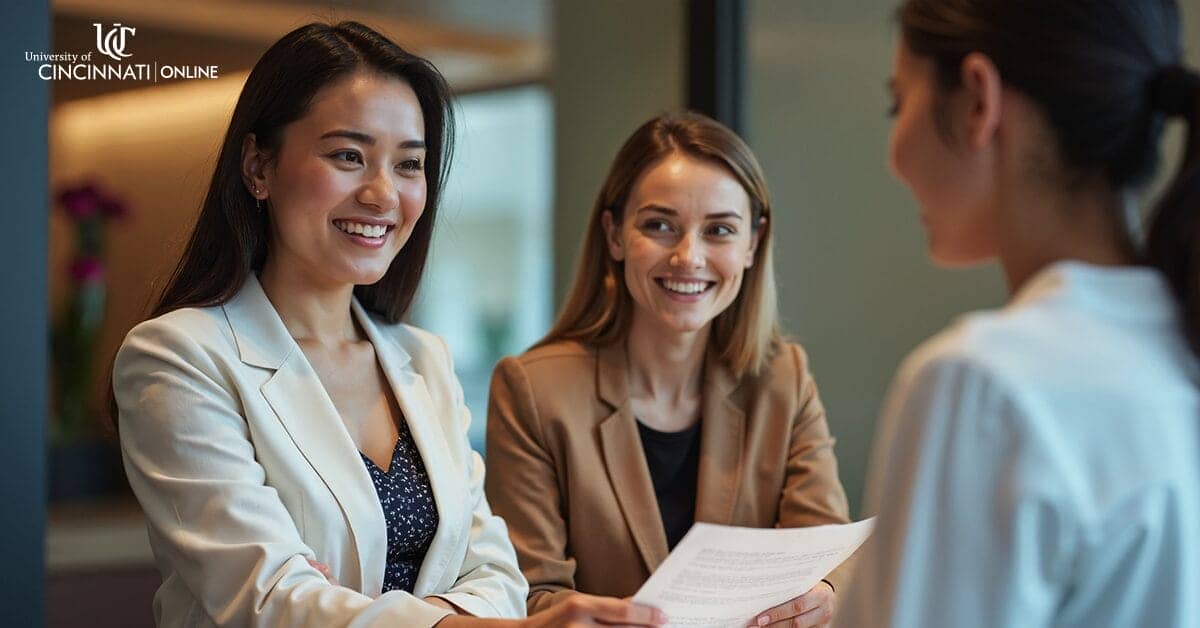 Two female hospitality management professionals smile at a guest who faces away from the viewer. One woman has long black hair and wears a white blazer. The other woman has light brown hair and wears a tan blazer, and she holds a sheet of paper.