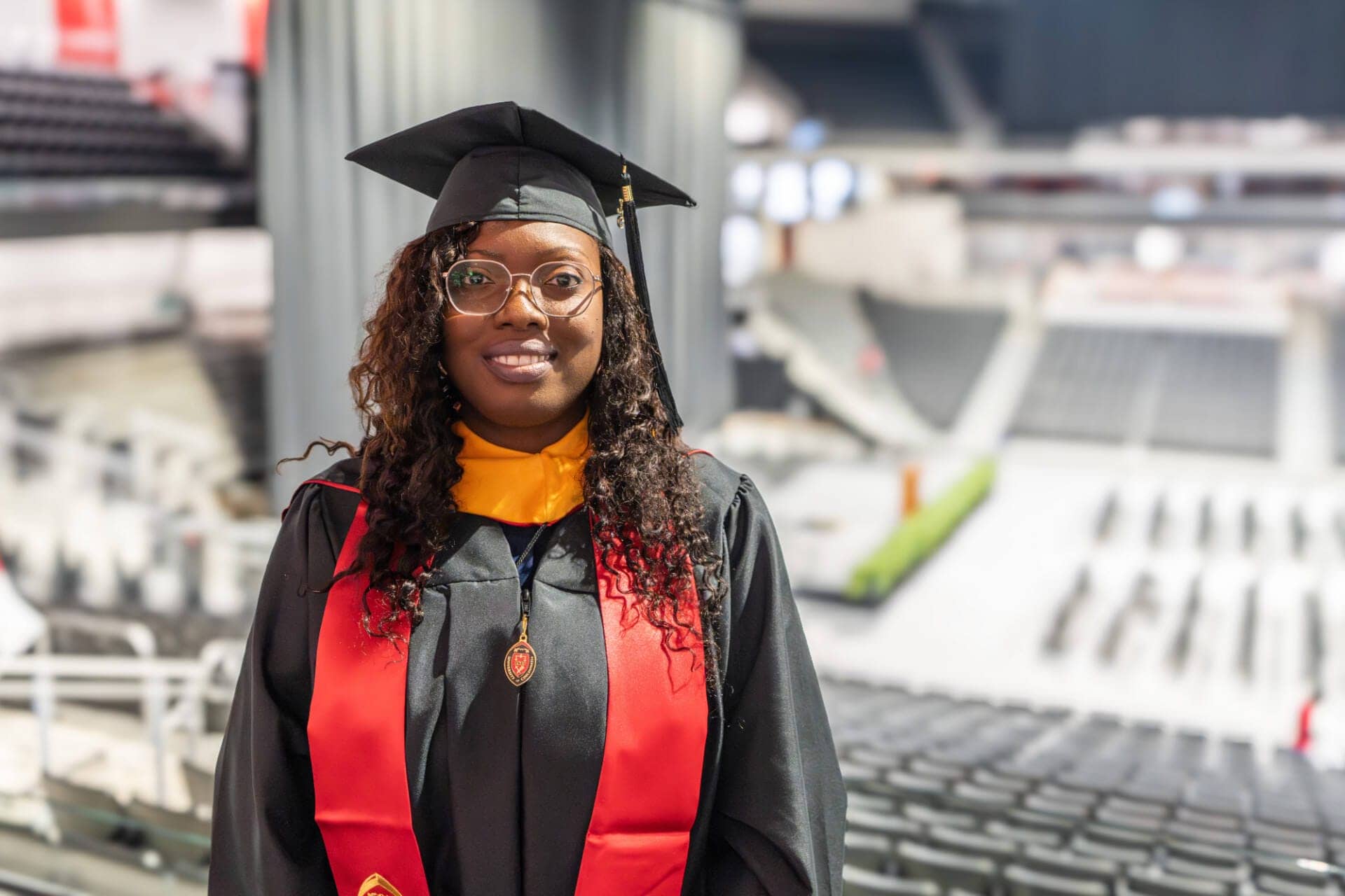 A woman stands in front of a stadium in a graduation cap, black graduation gown, and red stole. She smiles at the camera. 