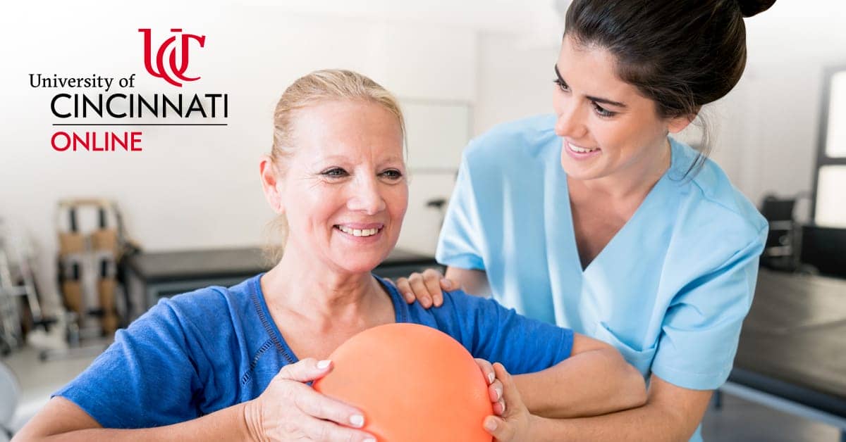 An occupational therapist rests her hand on a female patient's shoulder. The patient holds an orange exercise ball in front of her chest and smiles.