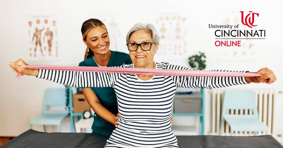 A female occupational therapist in green scrubs assists an adult client with occupational therapy exercises. The client, a woman with short grey hair, uses a pink elastic resistance band in front of her body to stretch.