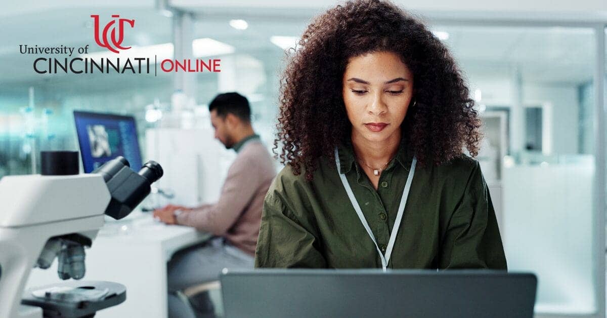A woman sits at a laptop in a lab. She is looking down and typing. To the left is a microscope and another man typing at a computer in the background.